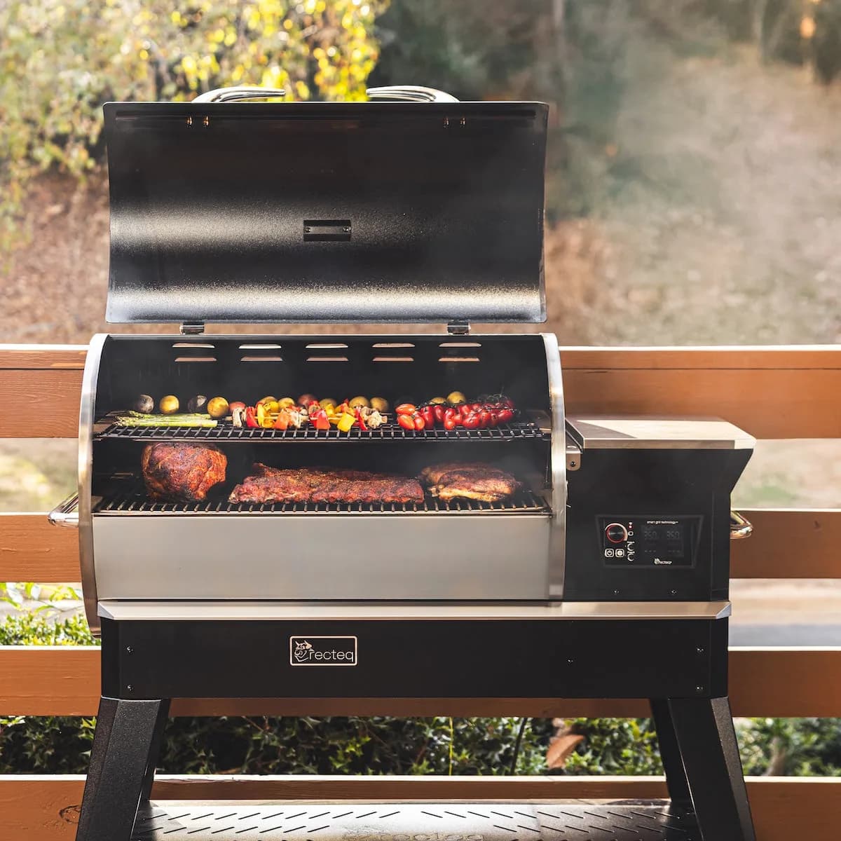 A Backyard Beast 1200 on a deck, lid open, grates loaded with ribs, sausages, and corn